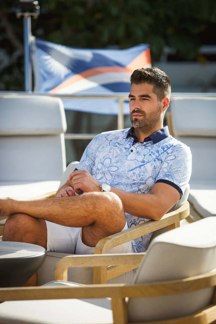 Homme portant un polo à motifs floraux, assis confortablement sur une chaise, décor extérieur élégant.