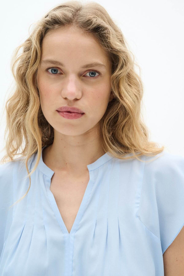 Blouse Cidenza légère en bleu clair avec encolure plissée et manches volantes, offrant élégance et confort.