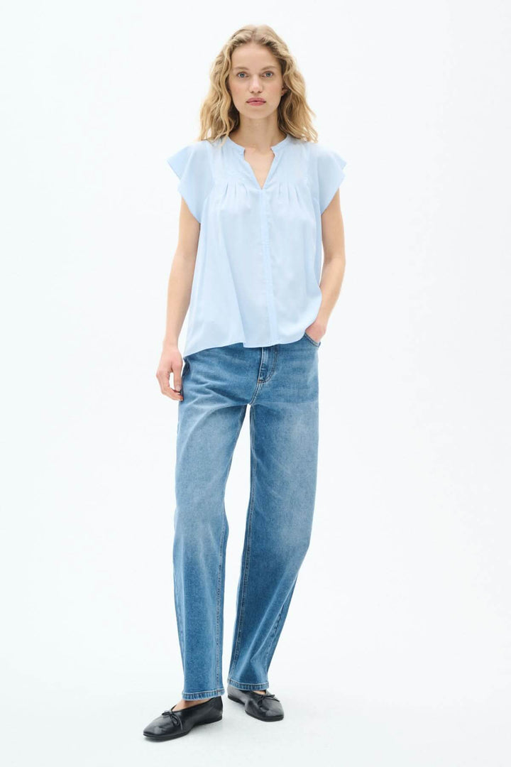Modèle en blouse légère bleue, manches volantes et coupe fluide, portée avec un jean et des chaussures noires.
