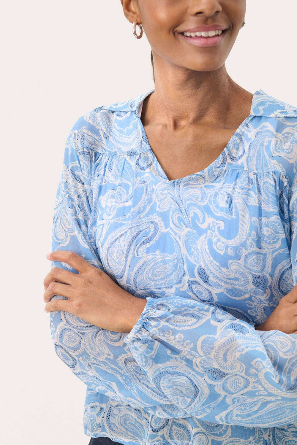Femme souriante portant une blouse légère en viscose avec un imprimé paisley bleu, encolure en V et manches bouffantes.