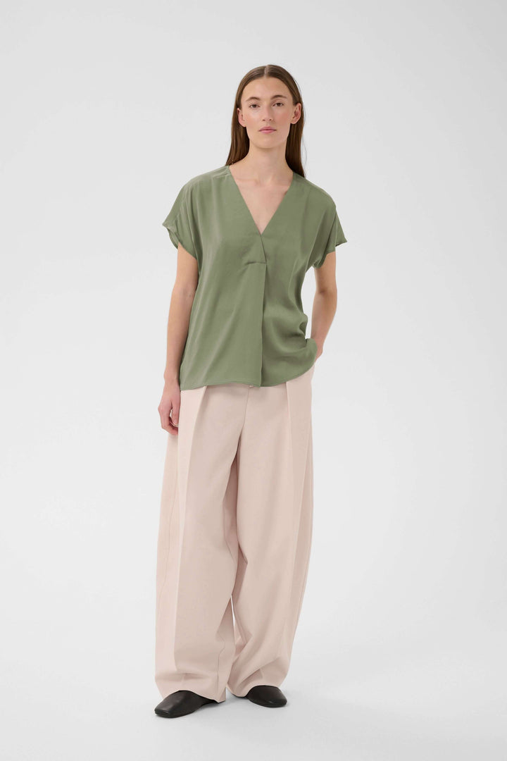Blouse Rinda fluide avec encolure en V, coupe droite et manches courtes, portée par une femme. Élégante et confortable pour le bureau.
