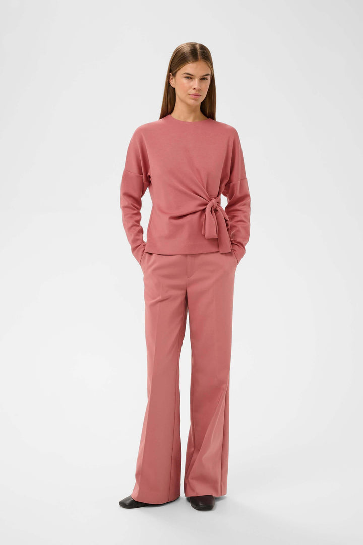 Femme portant la Blouse Tricent au design contemporain avec détail noué sur le côté, combinée avec un ensemble élégant et confortable.