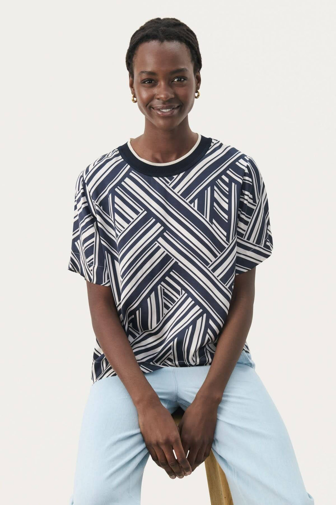 Femme souriante portant un tee-shirt à motifs géométriques navy et blanc, assise sur une chaise en bois.