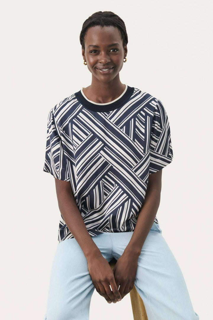 Femme souriante portant un tee-shirt à motifs géométriques navy et blanc, assise sur une chaise en bois.