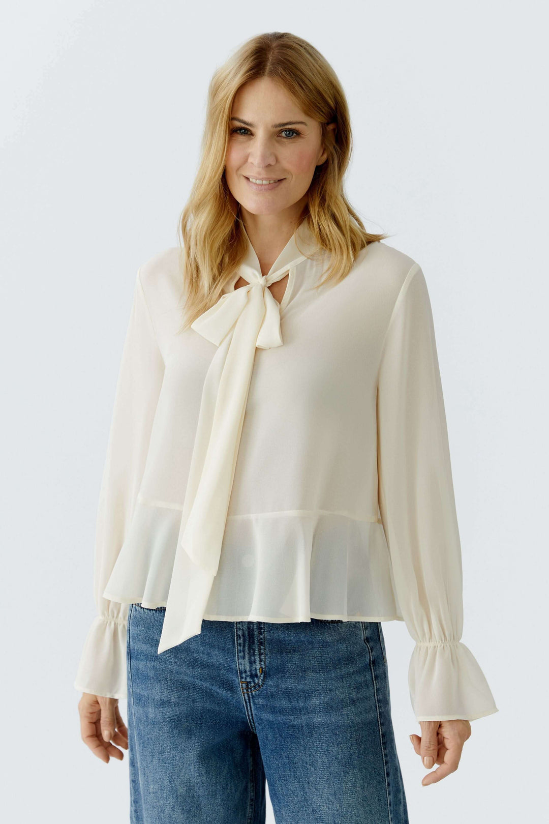 Blouse fluide à cravate col noué, élégante et féminine avec manches longues volantées, portée par une femme devant un fond neutre.