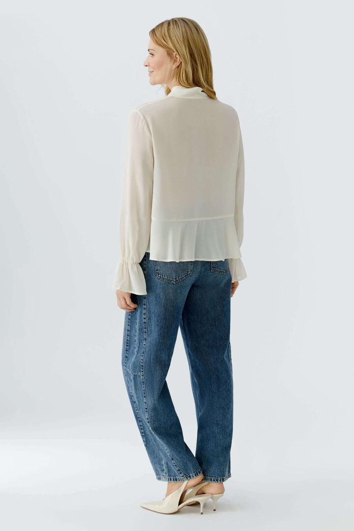 Femme portant une blouse fluide à cravate col noué, derrière, manches longues volantées, style élégant avec jean décontracté.