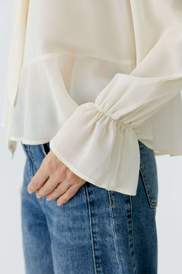 Blouse fluide à cravate col noué avec manches longues volantées, drapée sur un jean, élégance féminine. Vue rapprochée.