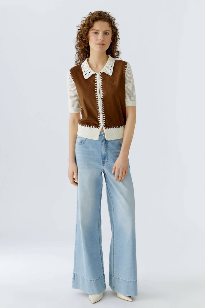 Femme portant une blouse manches courtes marron avec détails contrastants, mise en valeur par un col ajouré, jeans évasés et talons blancs.