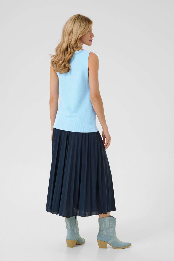 Femme portant un débardeur bleu clair et une jupe plissée noire, avec des bottes à talons turquoise.
