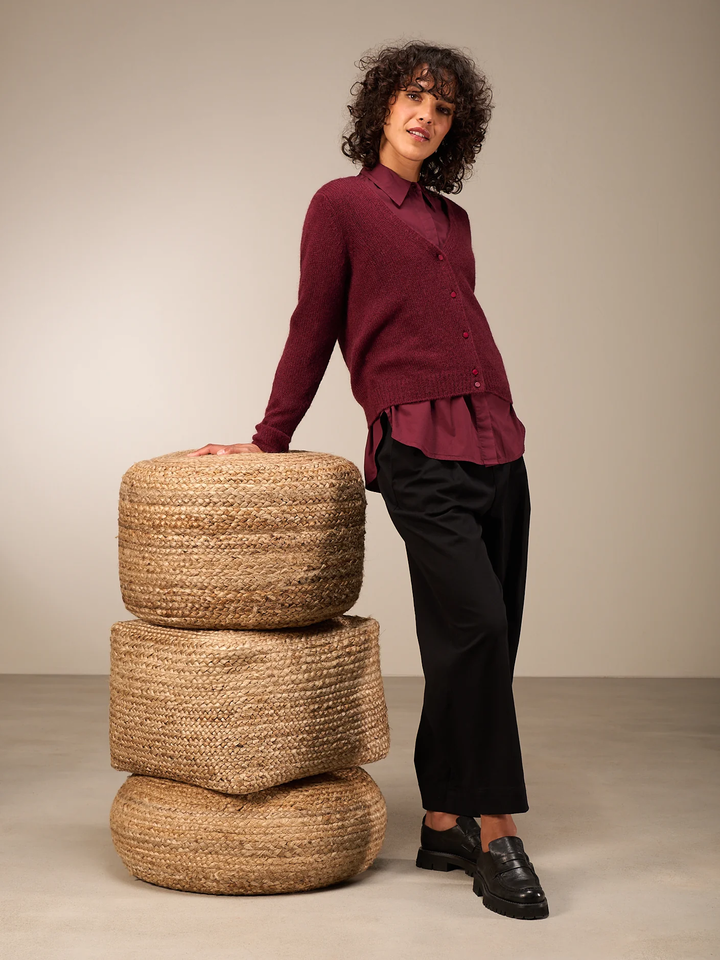 Femme portant un cardigan coupe droite bordeaux avec encolure en V, sur une chemise, et posant près de poufs en osier.