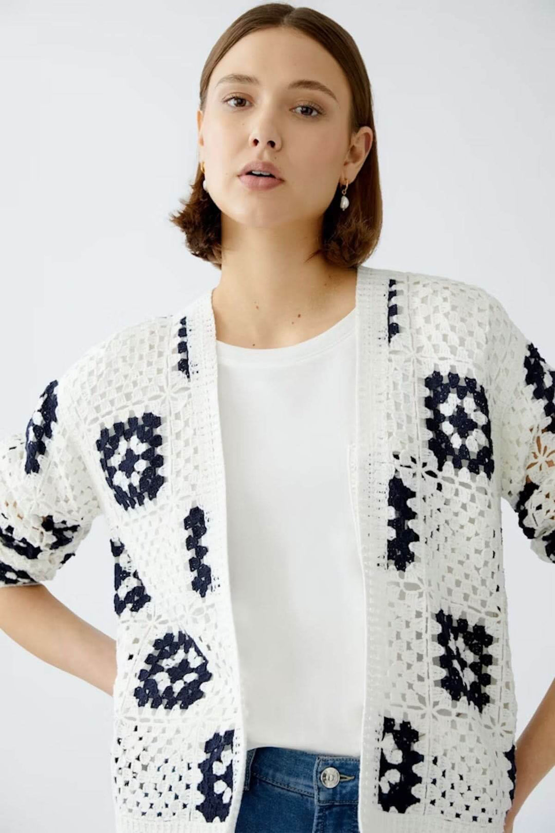 Cardigan en crochet blanc avec motifs géométriques noirs, porté sur un t-shirt blanc et un jean bleu.