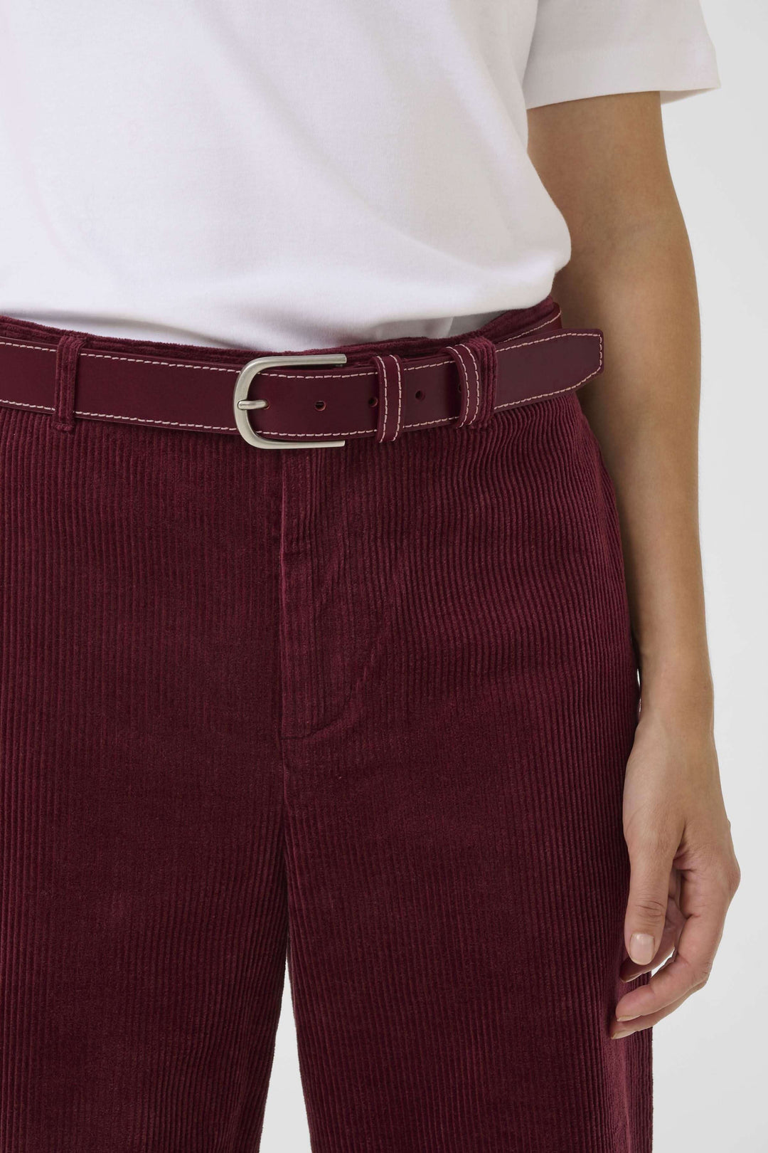 Ceinture Seyra en cuir véritable bordeaux avec surpiqûres contrastantes et boucle métallique, portée sur un pantalon en velours côtelé rouge.