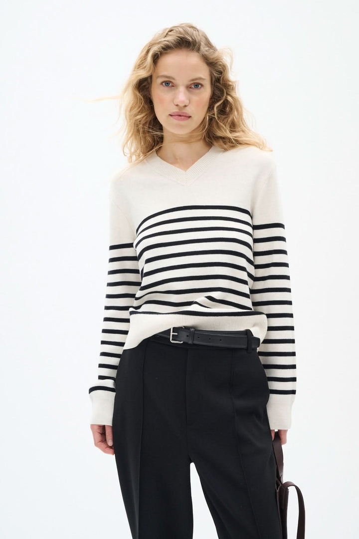Femme portant un pull beige à rayures noires avec un décolleté en V et un pantalon noir élégant.