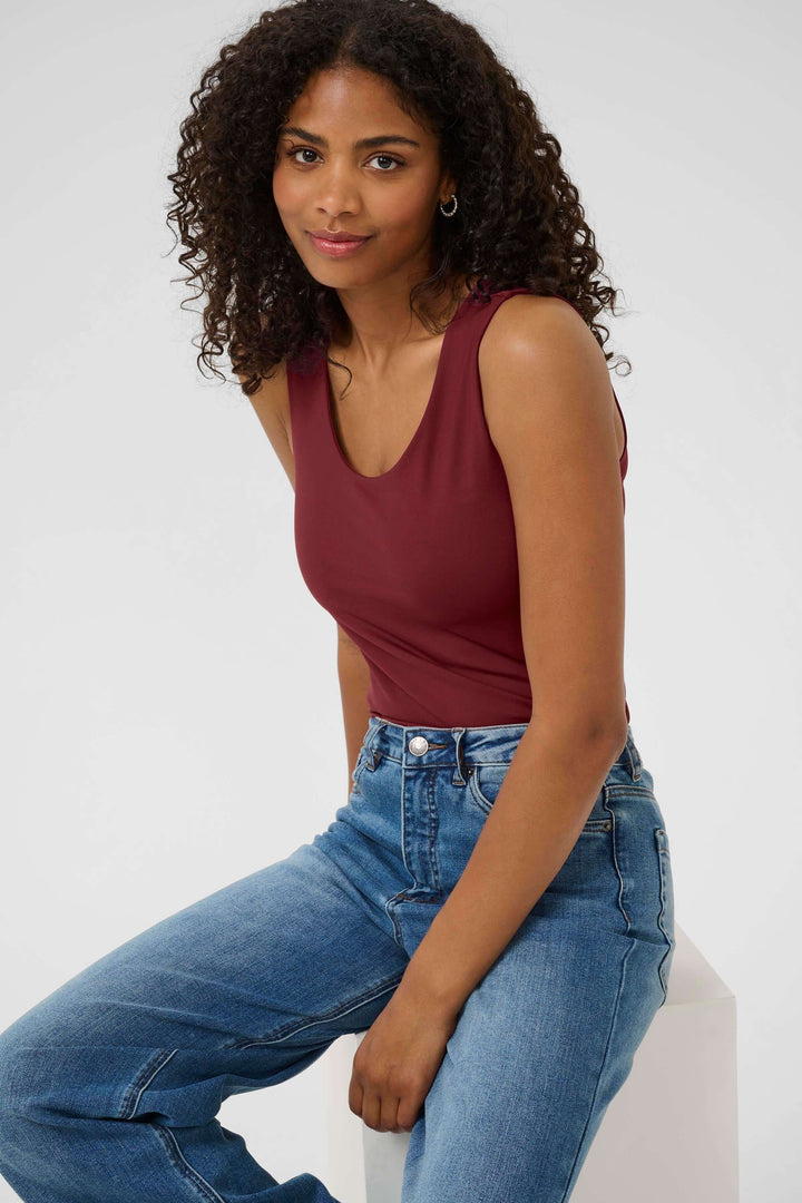 Femme portant un débardeur bordeaux simple avec jeans, assise sur un fond clair