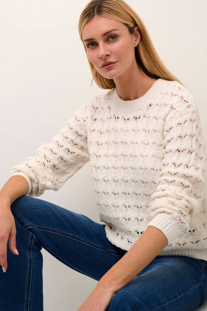 Femme portant un chandail Elena à manches longues, avec des détails en crochet et une coupe régulière, assise sur un sol.