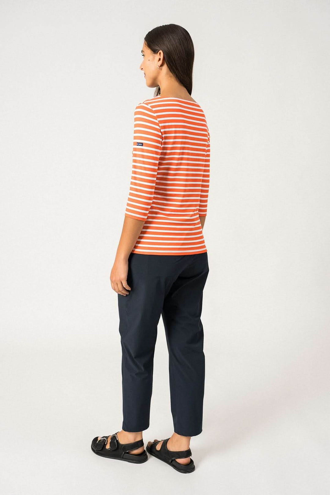 Femme de dos portant un chandail rayé orange et blanc, avec pantalon noir, montrant une coupe ajustée et moderne.