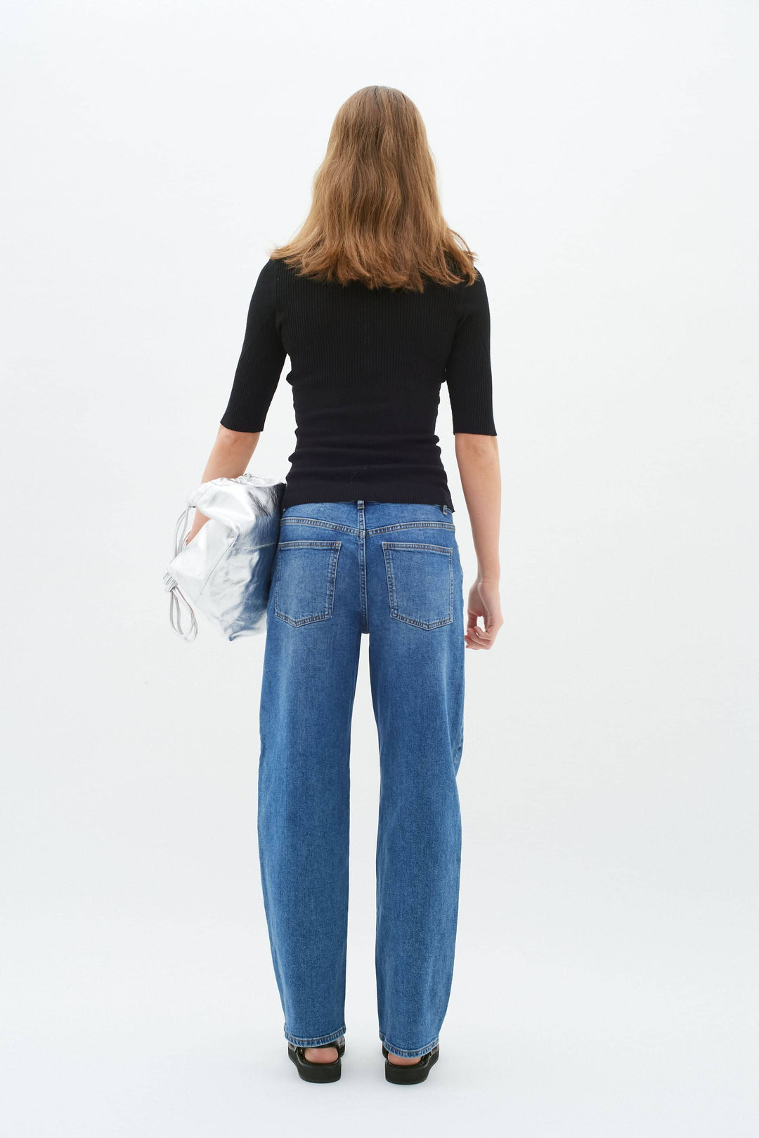 Femme vue de dos portant un chandail noir ajusté et un pantalon en denim bleu, tenant une veste argentée.