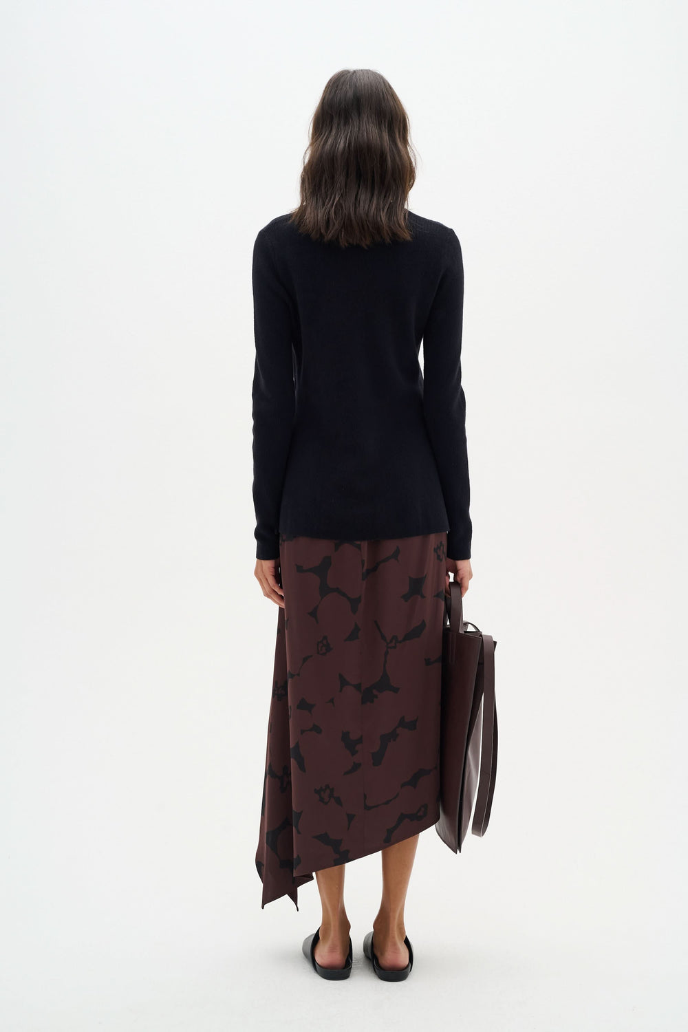 Femme de dos portant un chandail noir et une jupe marron avec un motif, tenant un sac marron.