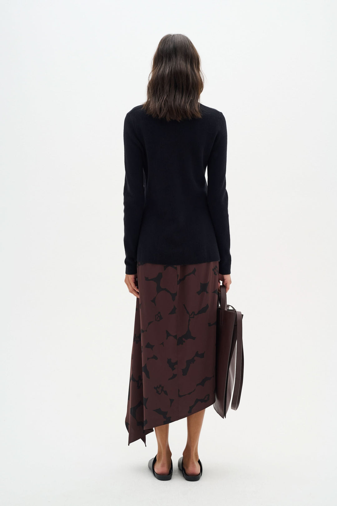 Femme de dos portant un chandail noir et une jupe marron avec un motif, tenant un sac marron.
