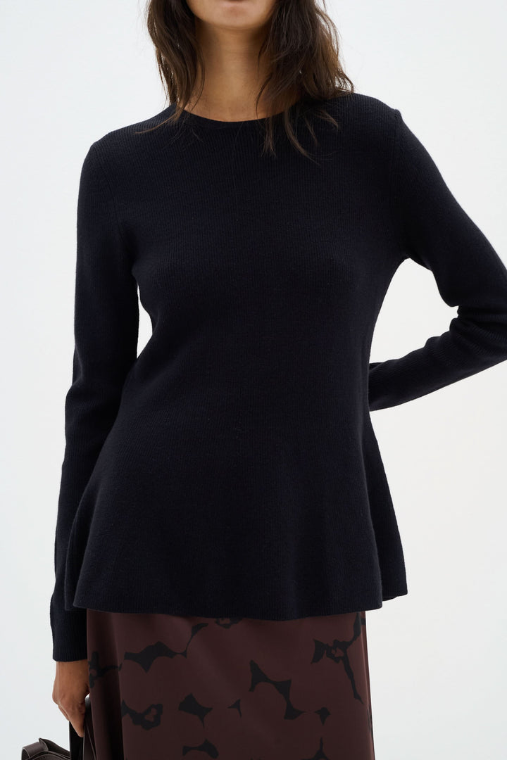 Chandail noir à col montant, coupe péplum élégante, porté avec une jupe, style sophistiqué.