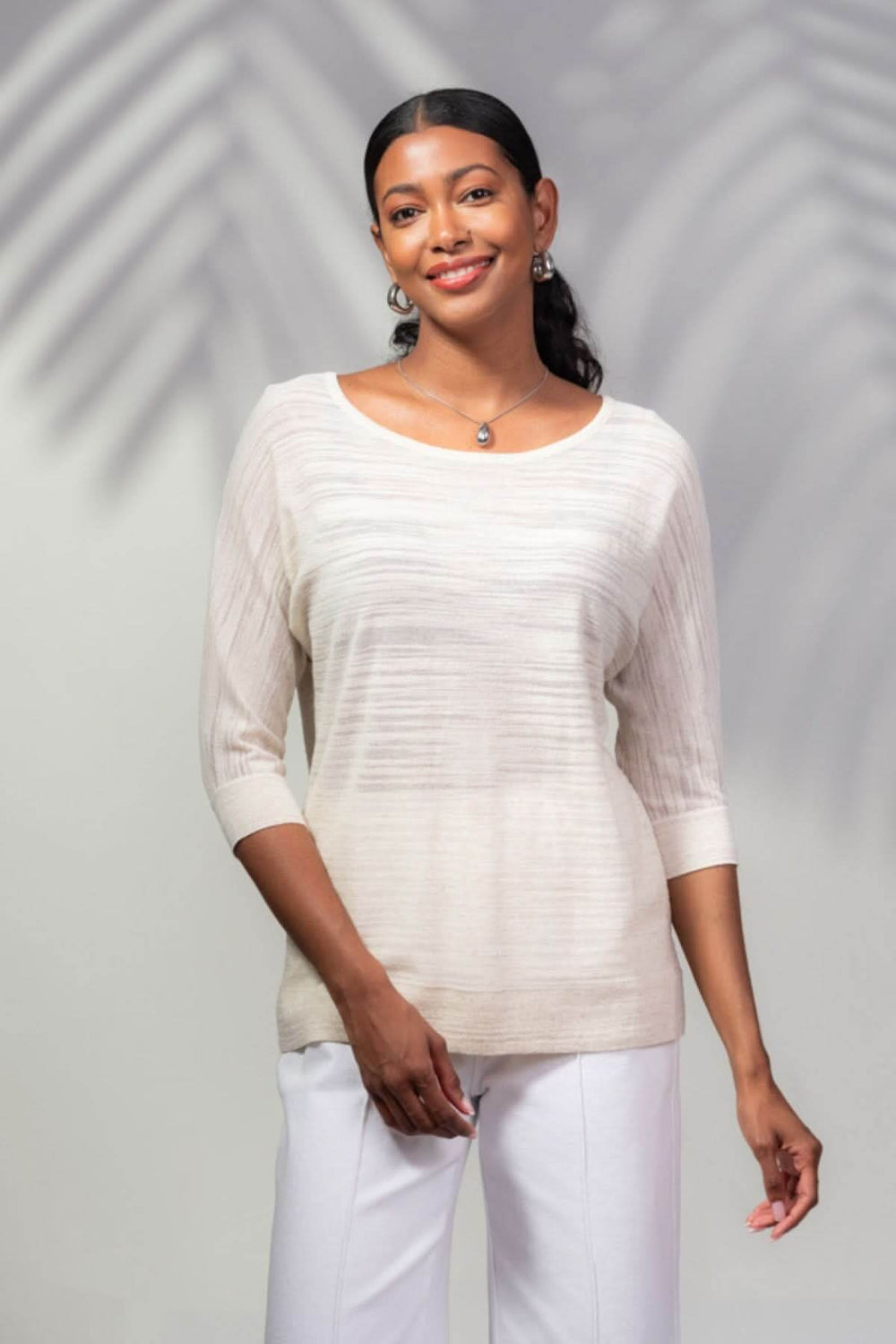 Chandail beige avec détails transparents, manches 3/4, encolure ronde, porté avec un pantalon blanc.