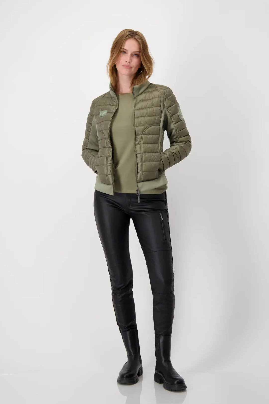 Femme portant un chandail basique vert sous une veste légère en olive, avec un pantalon noir en simili cuir.