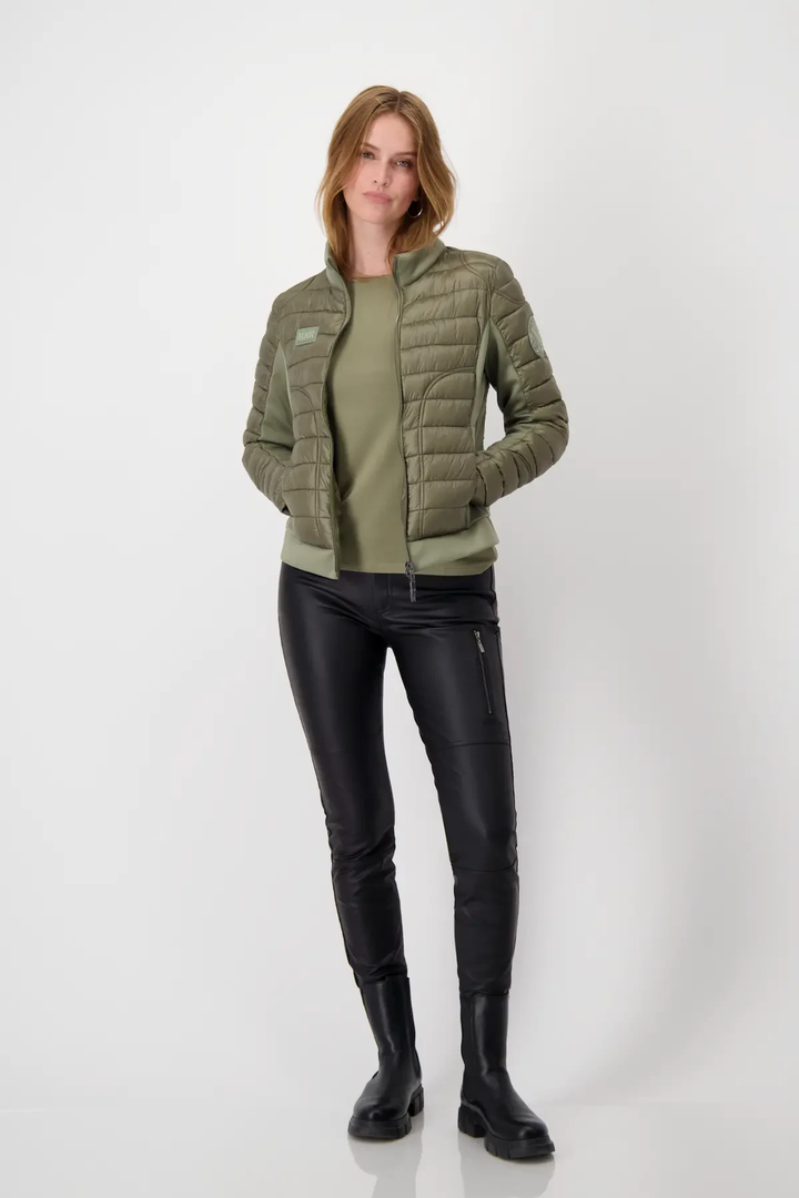 Femme portant un chandail basique vert sous une veste légère en olive, avec un pantalon noir en simili cuir.