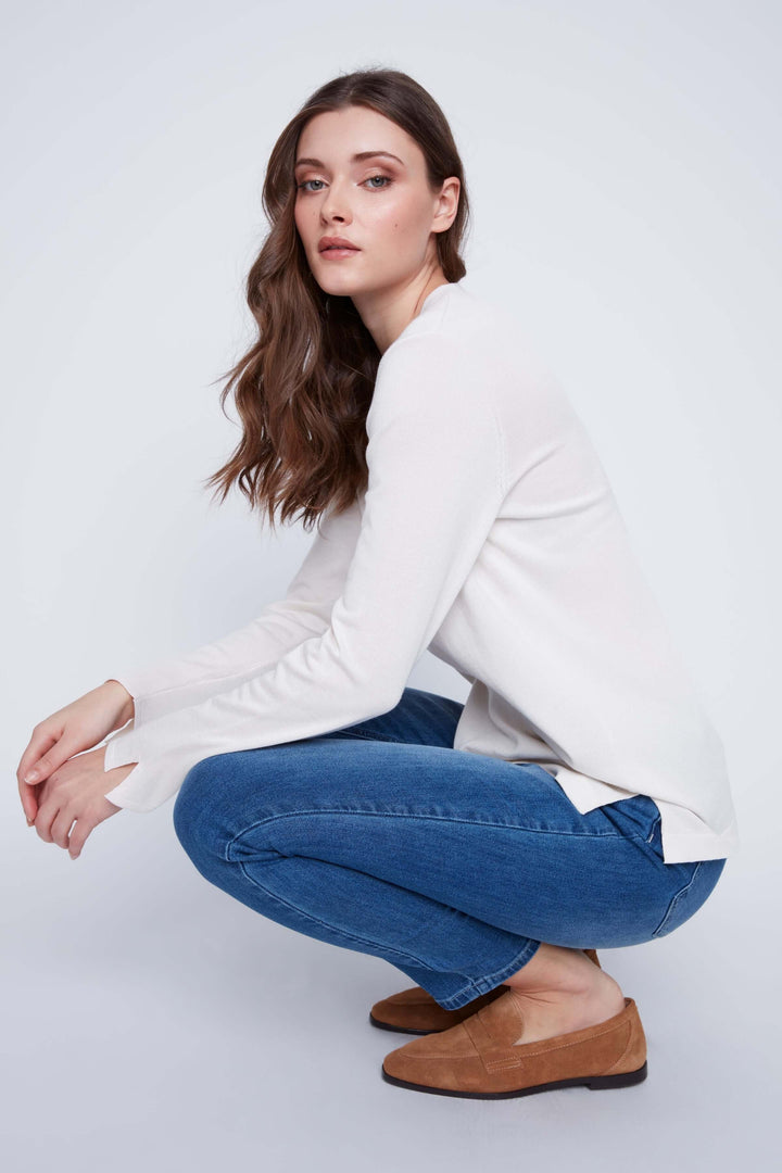 Femme portant un Chandail col V, couleur blanche, manches longues, style élégant et minimaliste, assise en tailleur, fond neutre.
