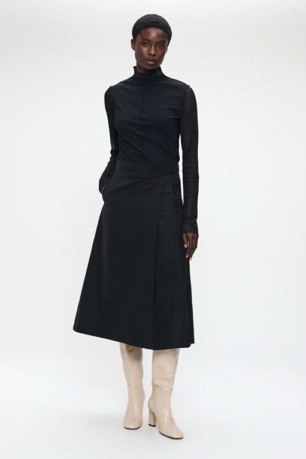 Femme portant un chandail à col montant et manches transparentes, élégant pour les occasions chics, avec une jupe noire et bottes blanches.