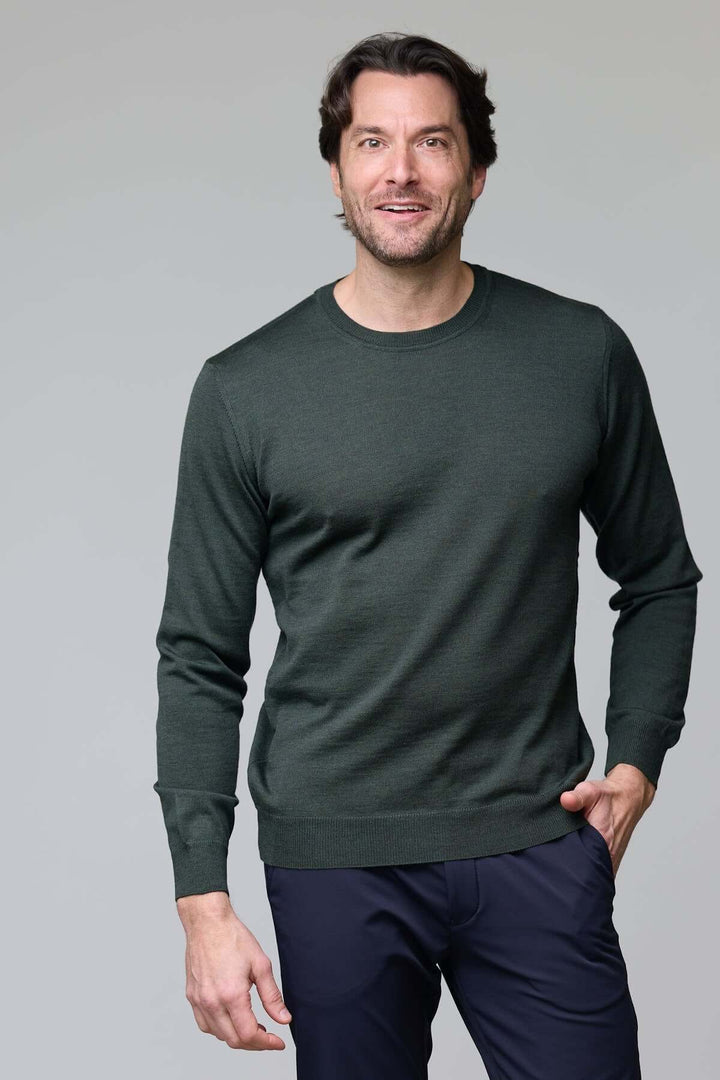 Homme portant un chandail vert à manches longues et pantalon bleu, posant avec un sourire.