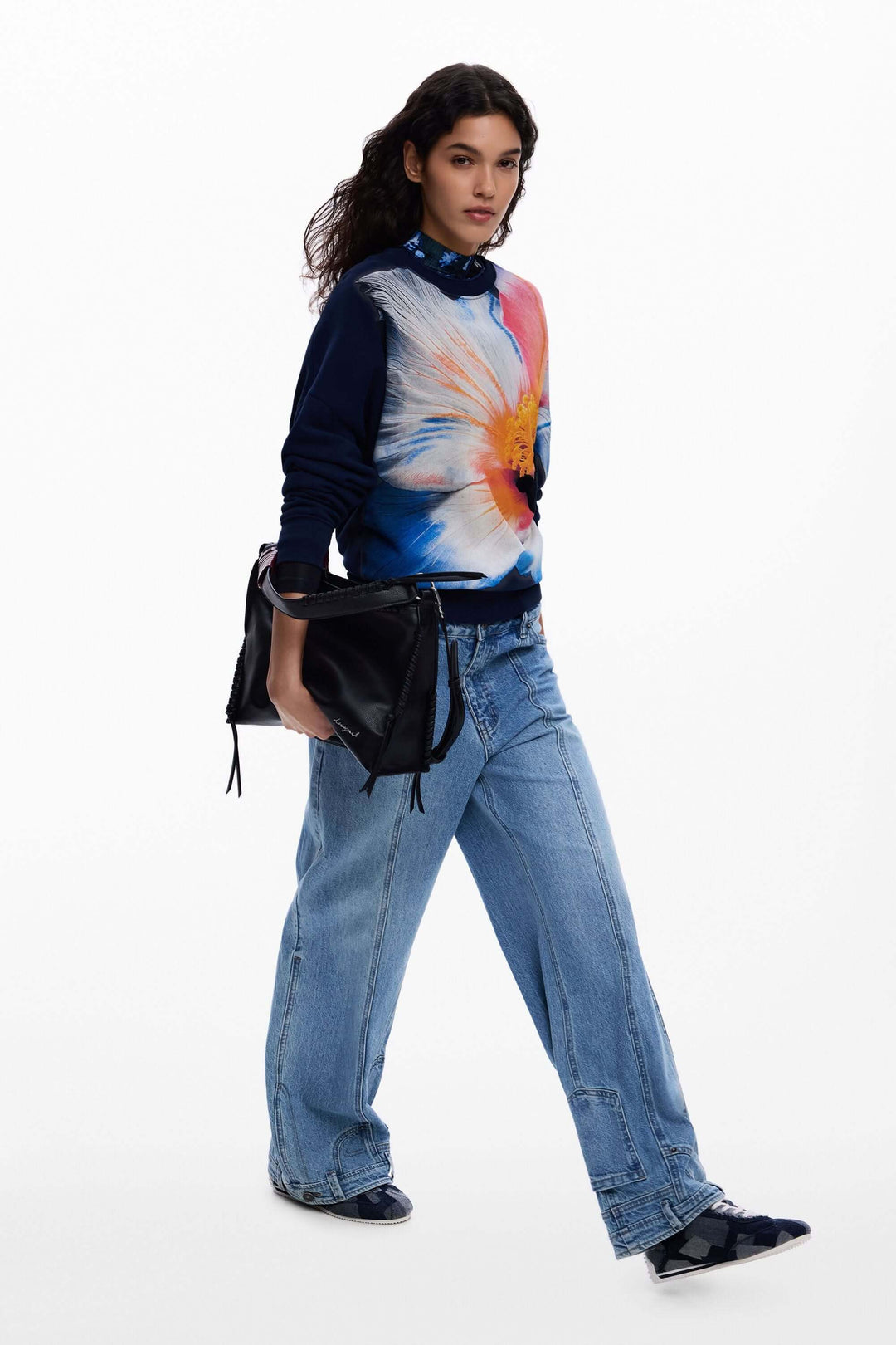 Femme portant un chandail à grande fleur avec un motif floral audacieux, assorti à un jean décontracté et une pochette noire.