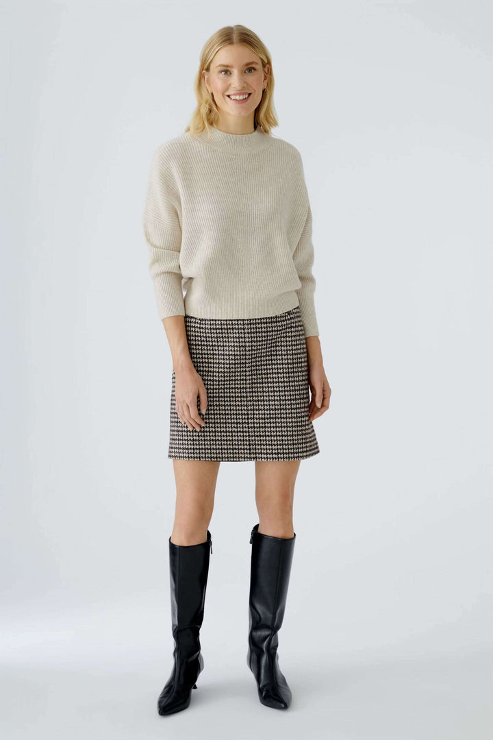 Chandail maille fine beige avec col montant, porté avec jupe à motifs et bottes noires, élégance minimaliste et confort moderne.