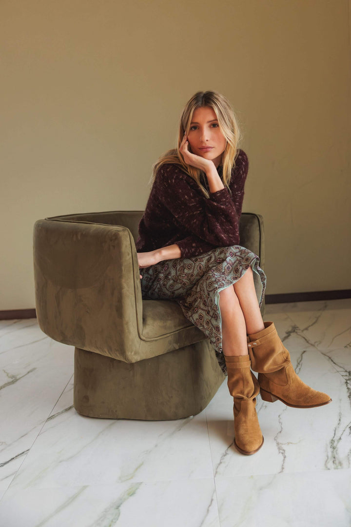 Femme assise portant un chandail maille texturée marron avec jupe imprimée et bottes camel dans un fauteuil moderne.
