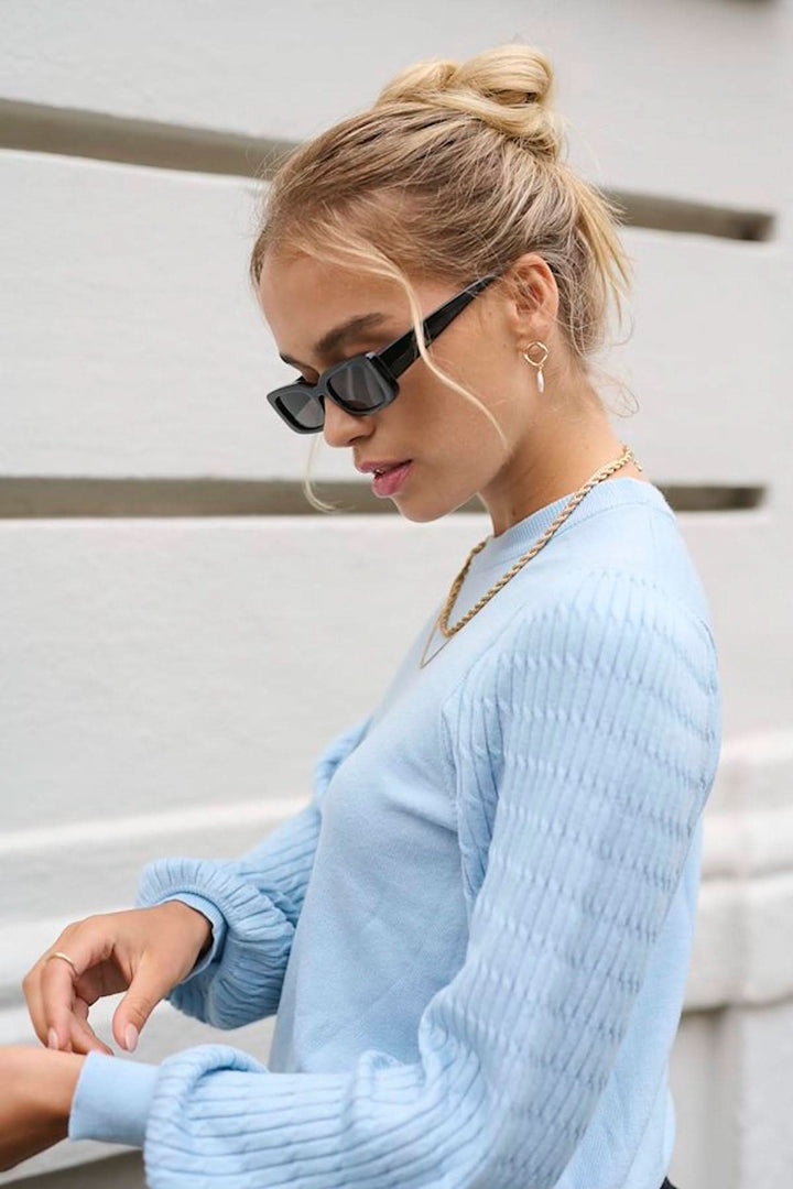 Femme portant un chandail bleu à manches à motifs et lunettes de soleil, montrant un style décontracté et moderne.