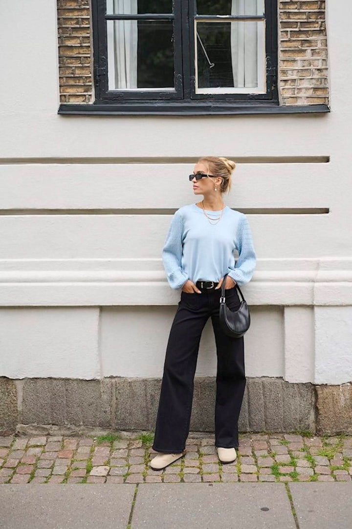 Femme portant un chandail bleu à manches amples et un pantalon noir, devant une fenêtre, style décontracté. 