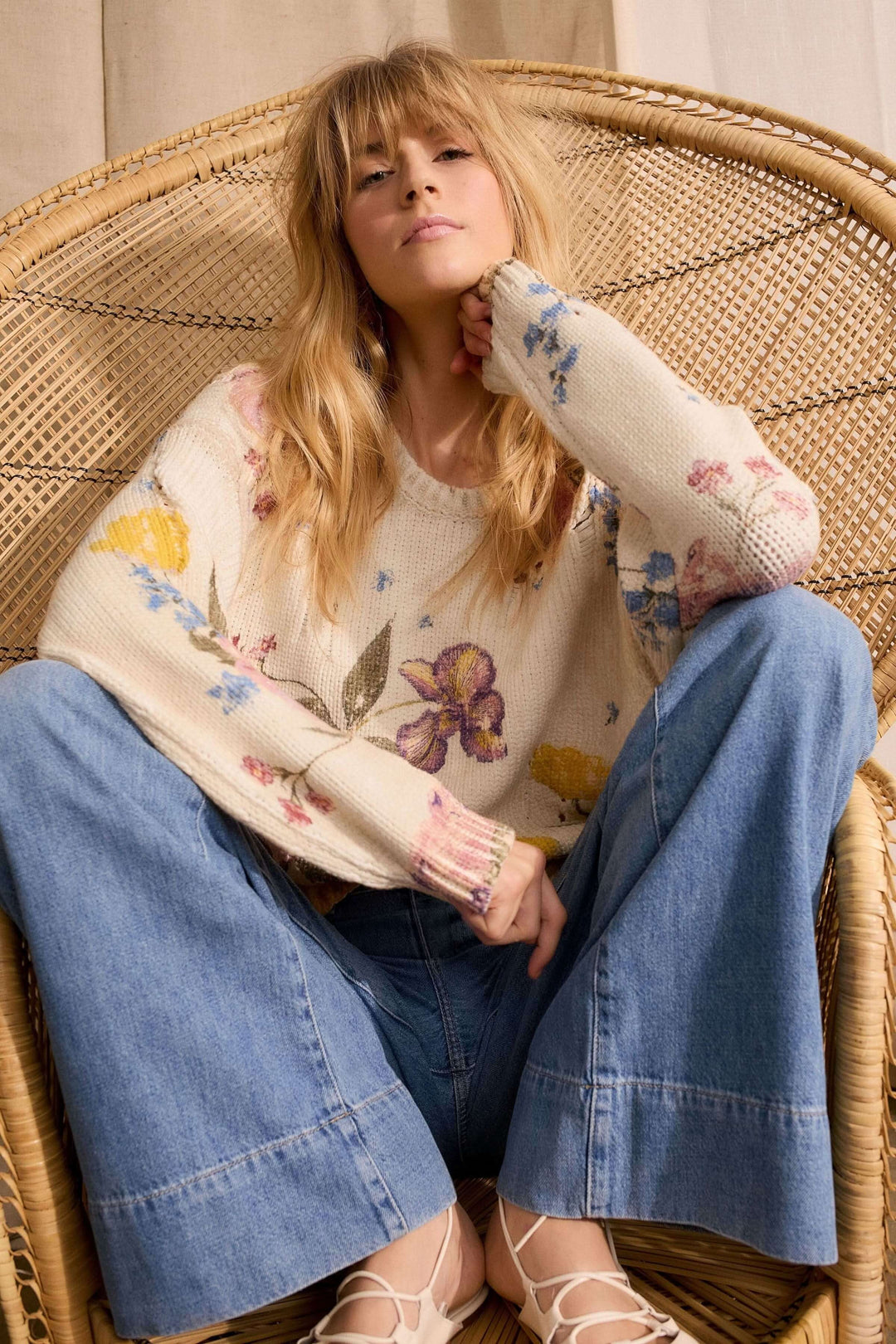 Femme portant un chandail vintage tendance avec imprimé floral et une coupe décontractée, assise sur une chaise en osier.