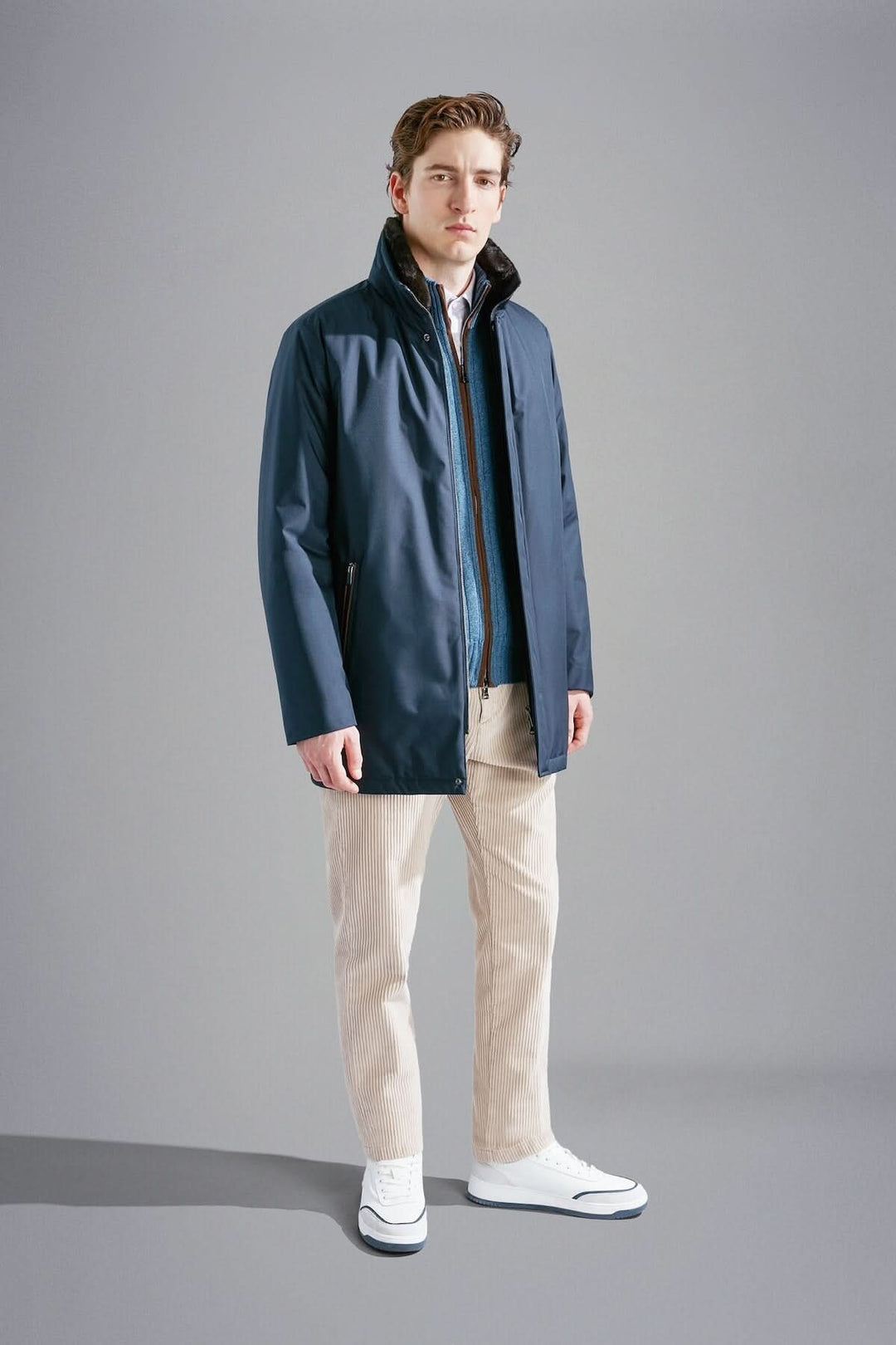 Homme en manteau bleu marine moderne, pantalon beige et baskets blanches sur fond gris.