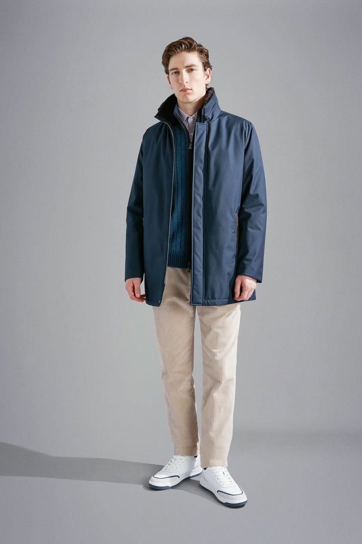Homme portant une veste bleue marine, pantalon beige et chaussures blanches dans un décor simple.