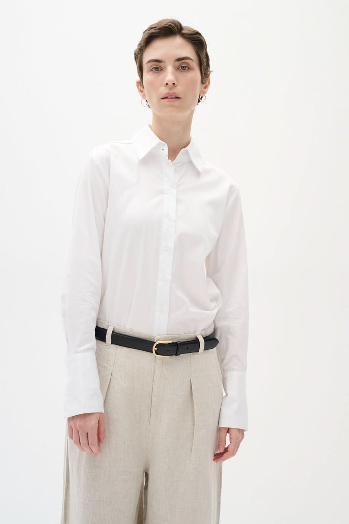 Chemise classique en coton blanche avec col traditionnel, portée avec un pantalon clair et une ceinture noire.