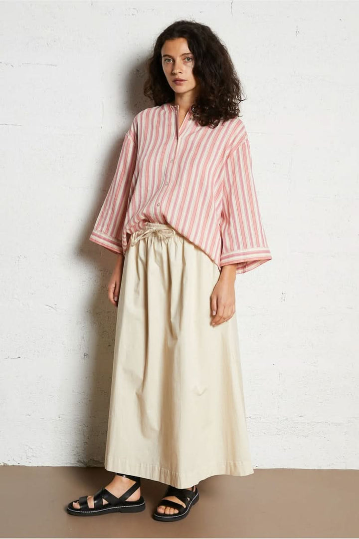 Chemise design épuré rayée rose avec coupe fluide, portée avec une jupe beige longue et des sandales noires. Décris moi le produit en français.