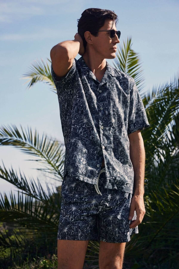 Homme en chemise et short à imprimé tropical, debout dans un environnement ensoleillé, entouré de palmiers.