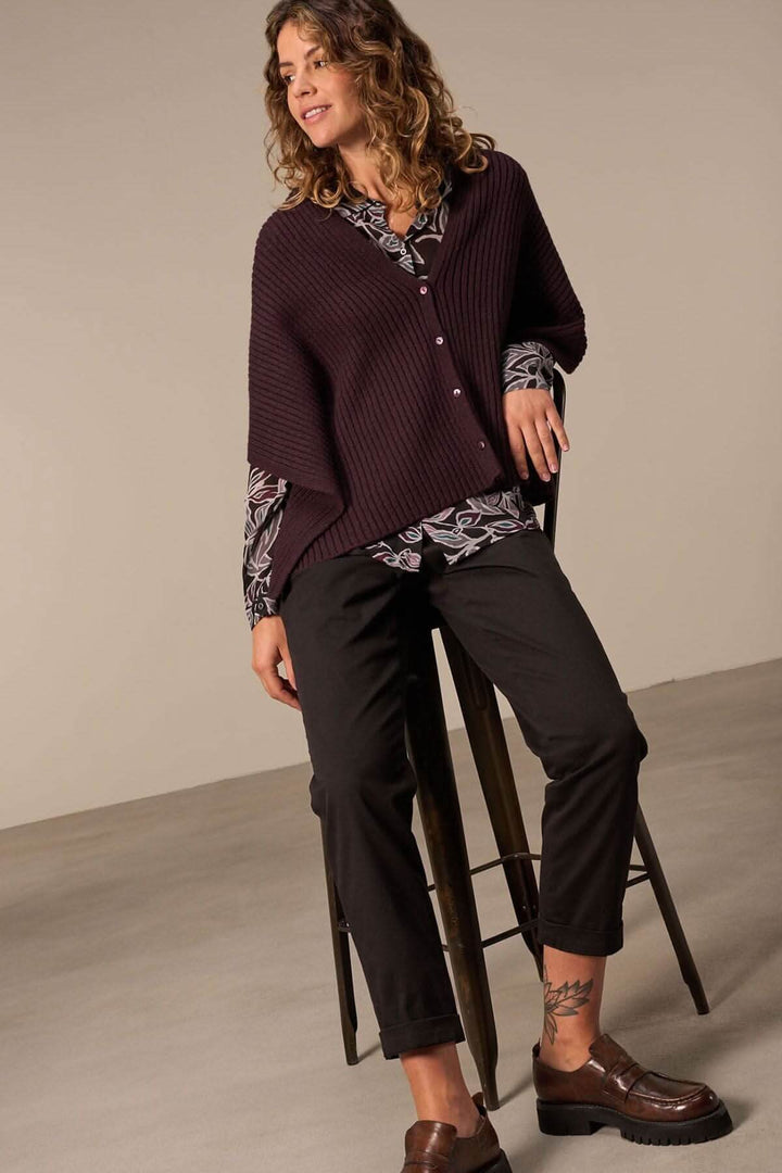 Femme en pull bordeaux et chemise à motifs, assise sur un tabouret avec un look élégant et décontracté.