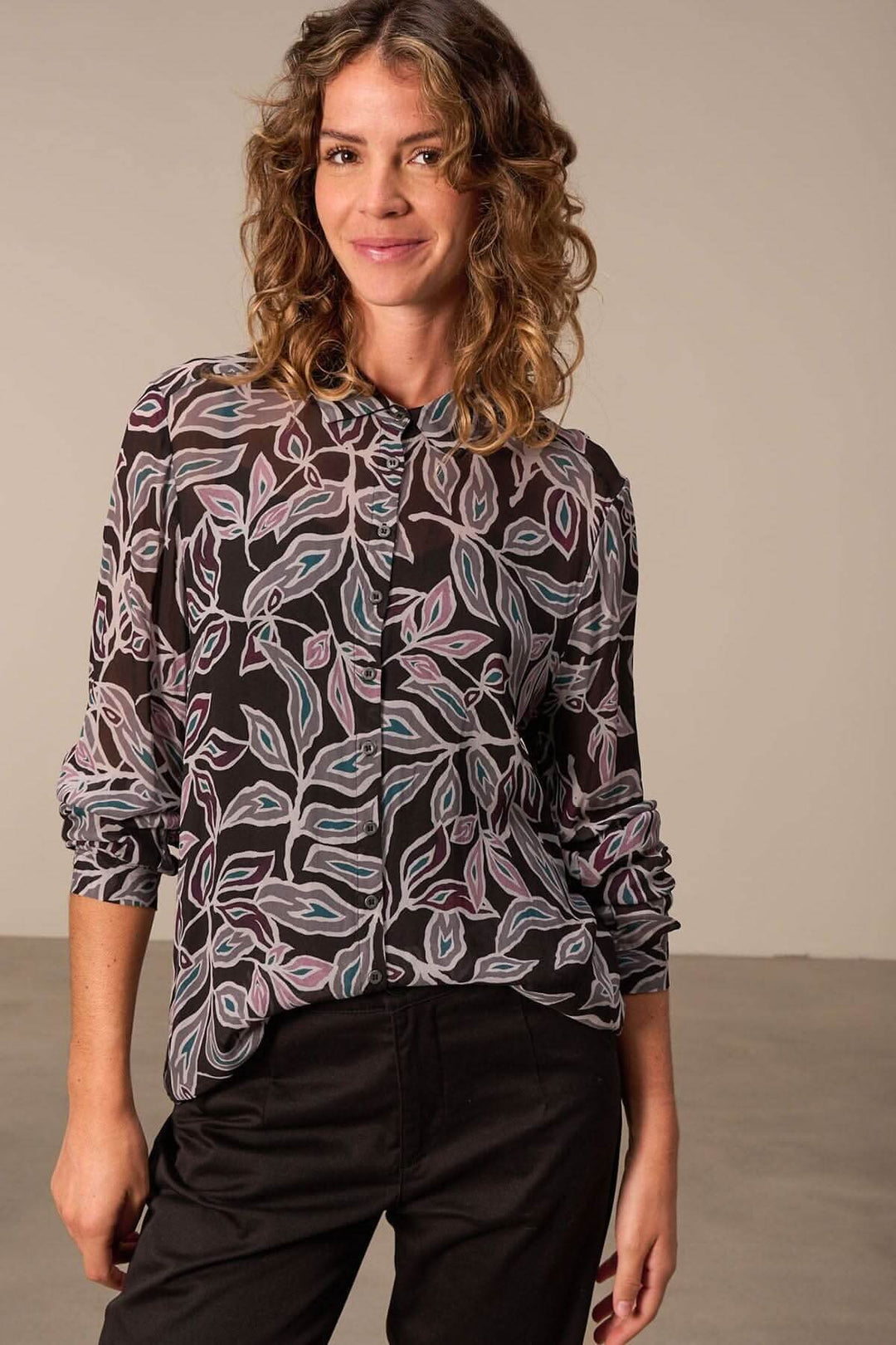Chemise semi transparente à imprimé floral, manches longues, portée par une femme souriante avec un style décontracté-chic.