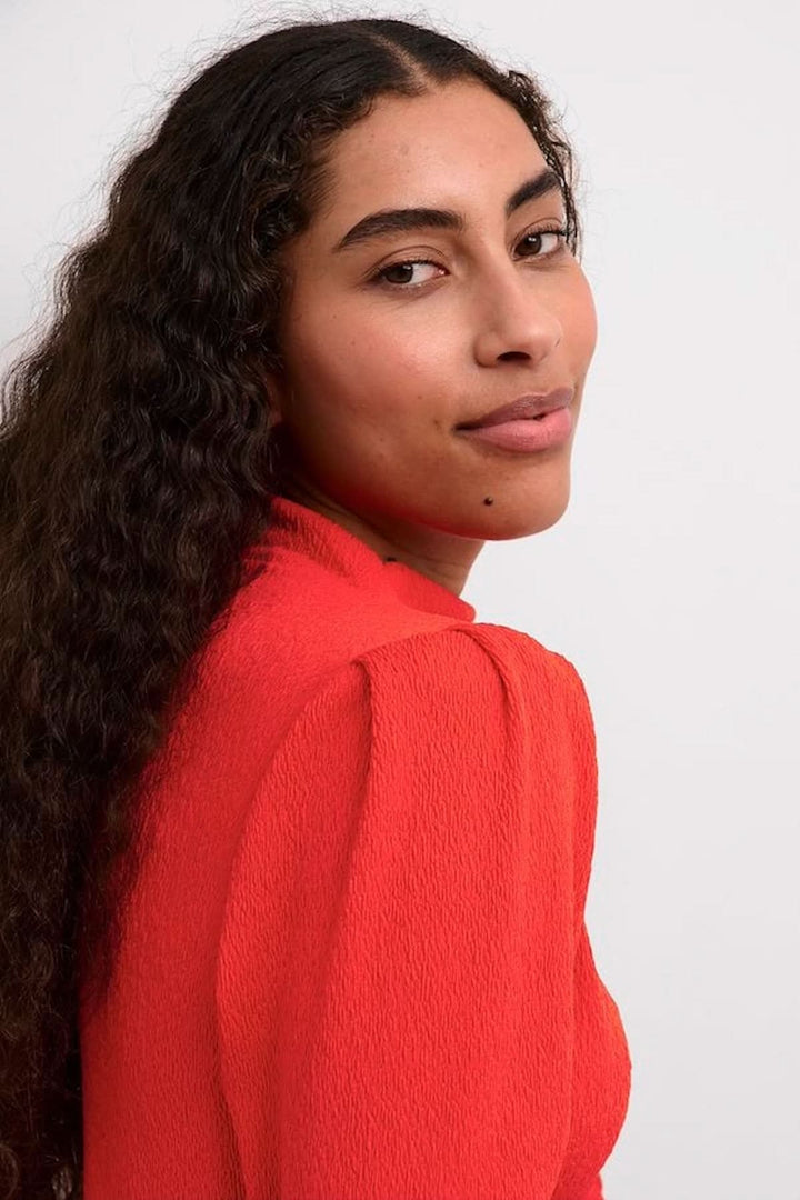 Femme avec cheveux bouclés portant un chemisier rouge vif à manches longues, vue de profil.