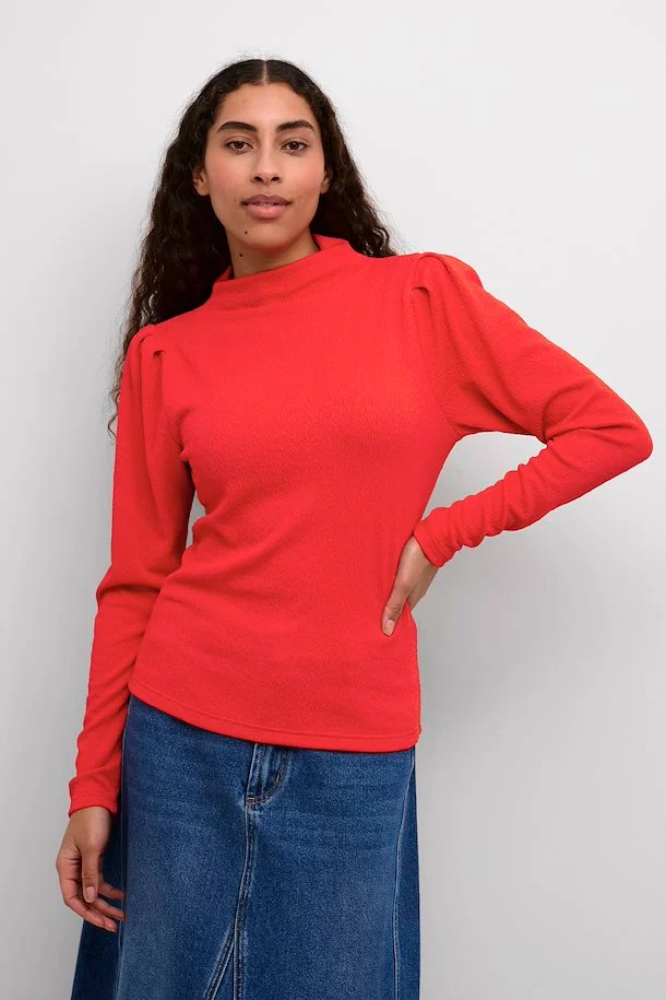 Chemisier rouge vif à manches longues et col rond, design structuré aux épaules, porté avec jupe en denim.