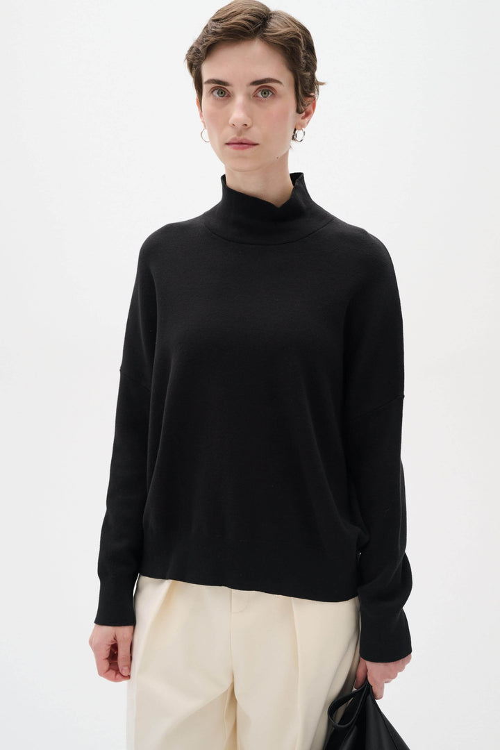 Chandail en coton noir à col roulé, manches longues, coupe ample et stylée, porté avec un pantalon crème.