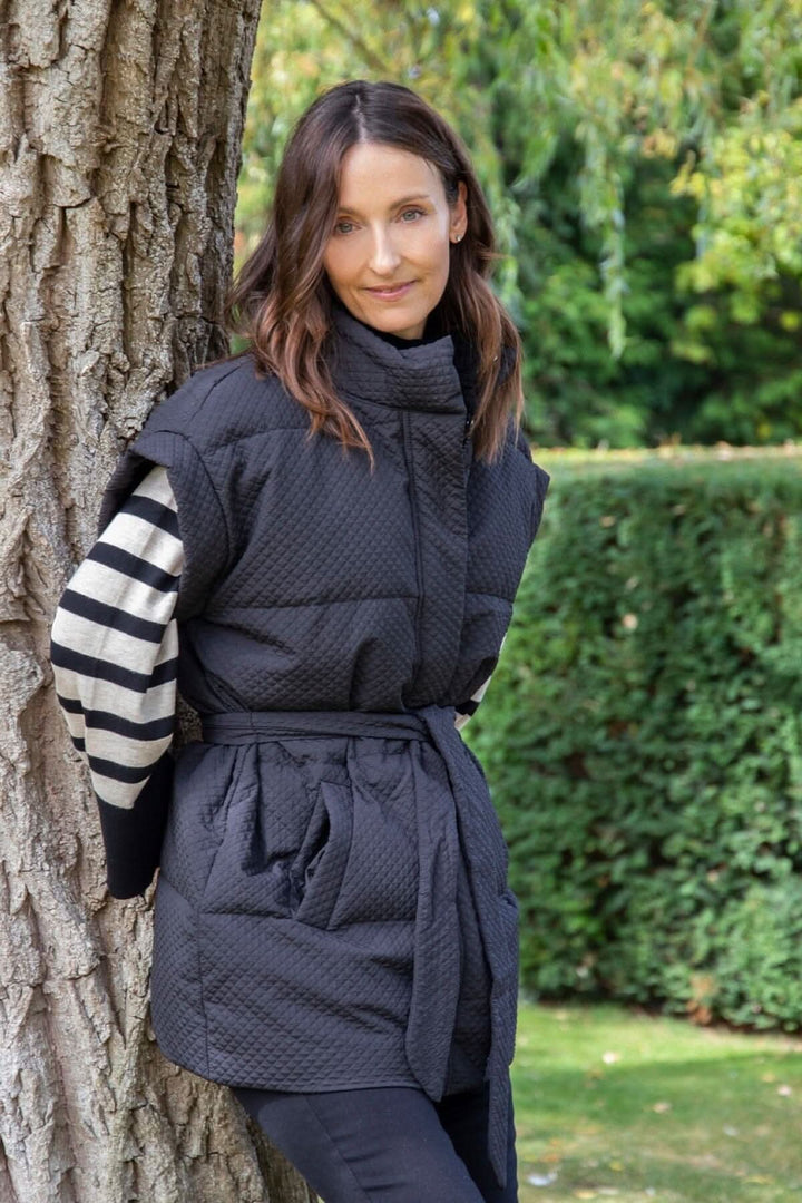 Modèle portant un gilet matelassé noir Tahani avec ceinture, posé contre un arbre, entouré d&