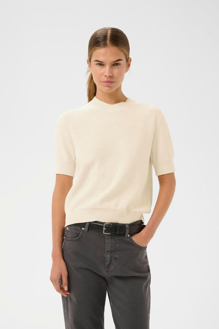 Haut à manches courtes NAASU en maille blanche, style épuré et raffiné, avec encolure arrondie, modèle féminin portant le haut seul.