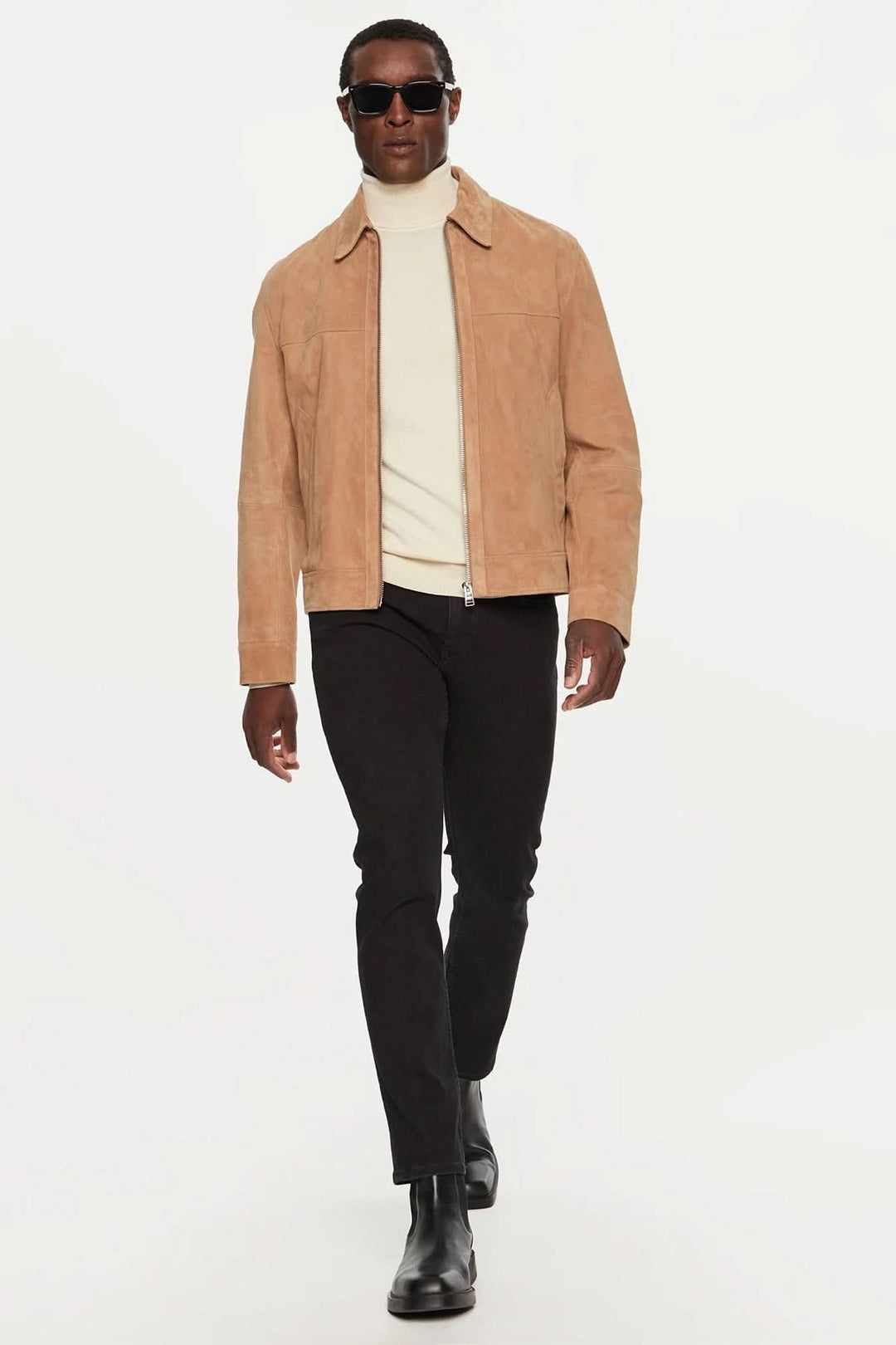 Homme marchant avec une veste en daim beige, pull blanc et jeans noirs élégants.