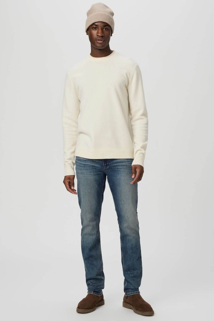 Homme portant un pull crème, un jean bleu délavé et un bonnet beige pour un look décontracté.